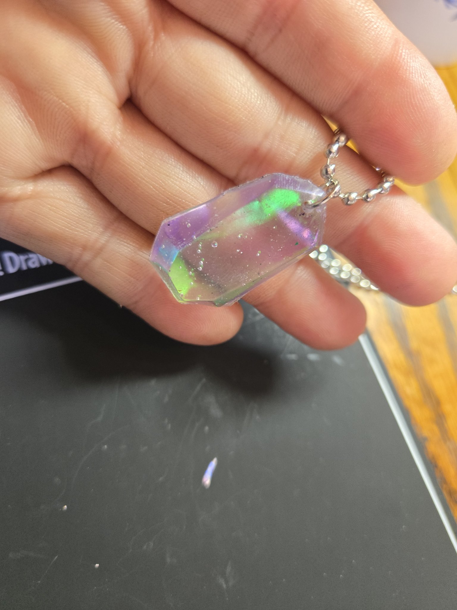 Holographic resin crystal pendant held in a hand, showing its translucent green and purple color shift