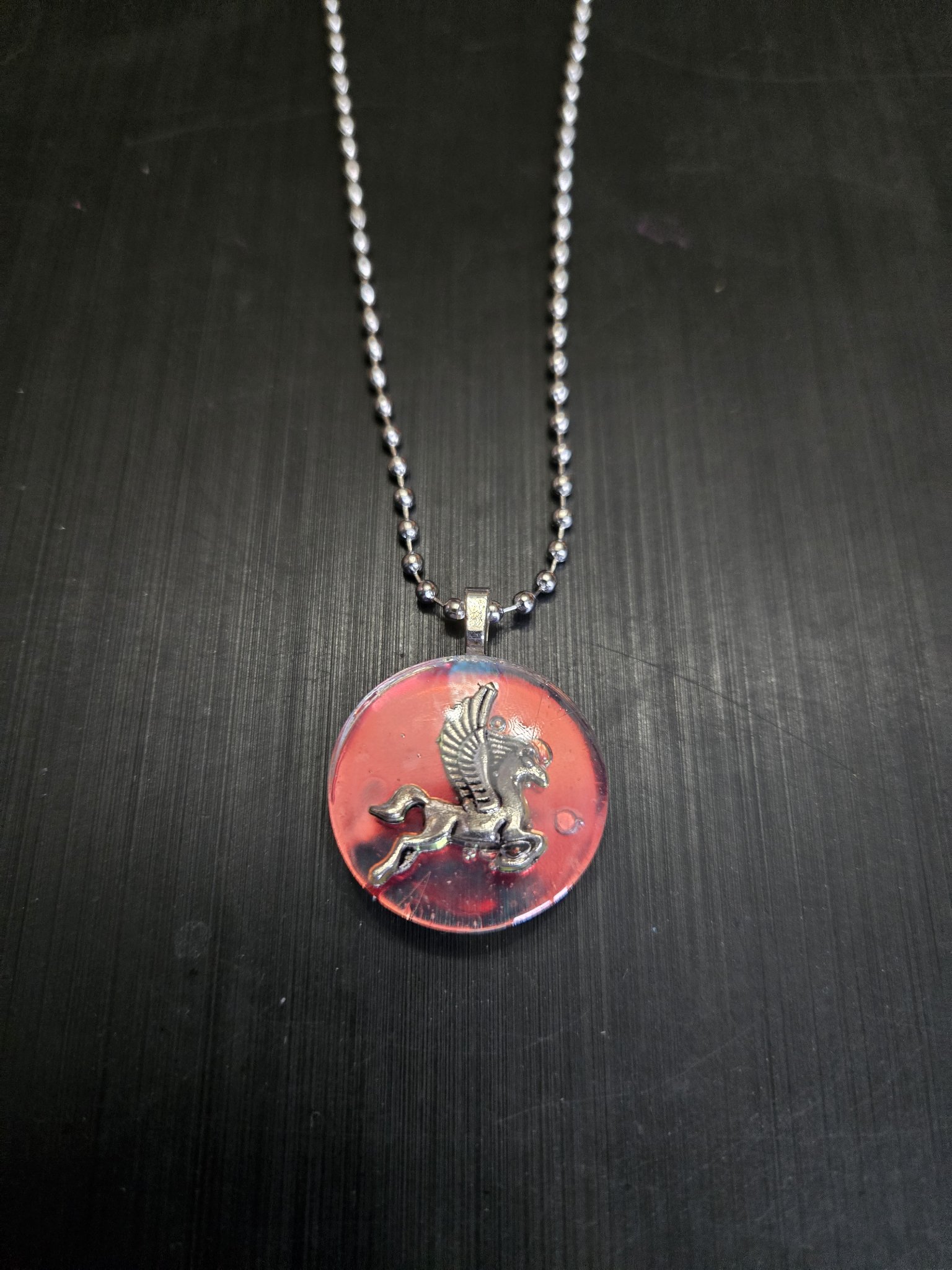 Front view of a round red and pink holographic resin pendant with a silver-tone pegasus charm on a ball chain