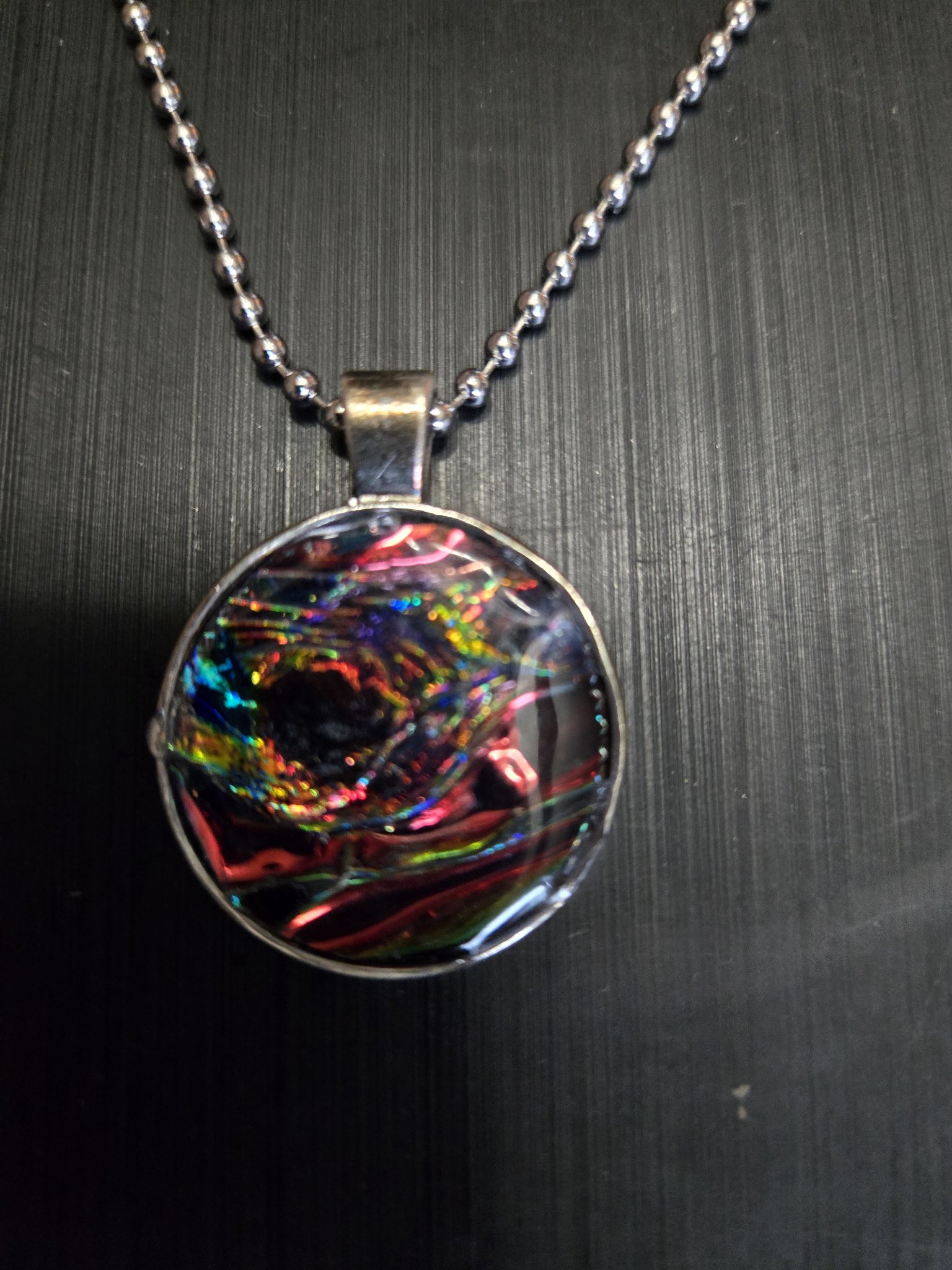 Front view of the round metal and resin holographic pendant with a dark red, green, and blue rainbow swirl on its 22-inch ball chain