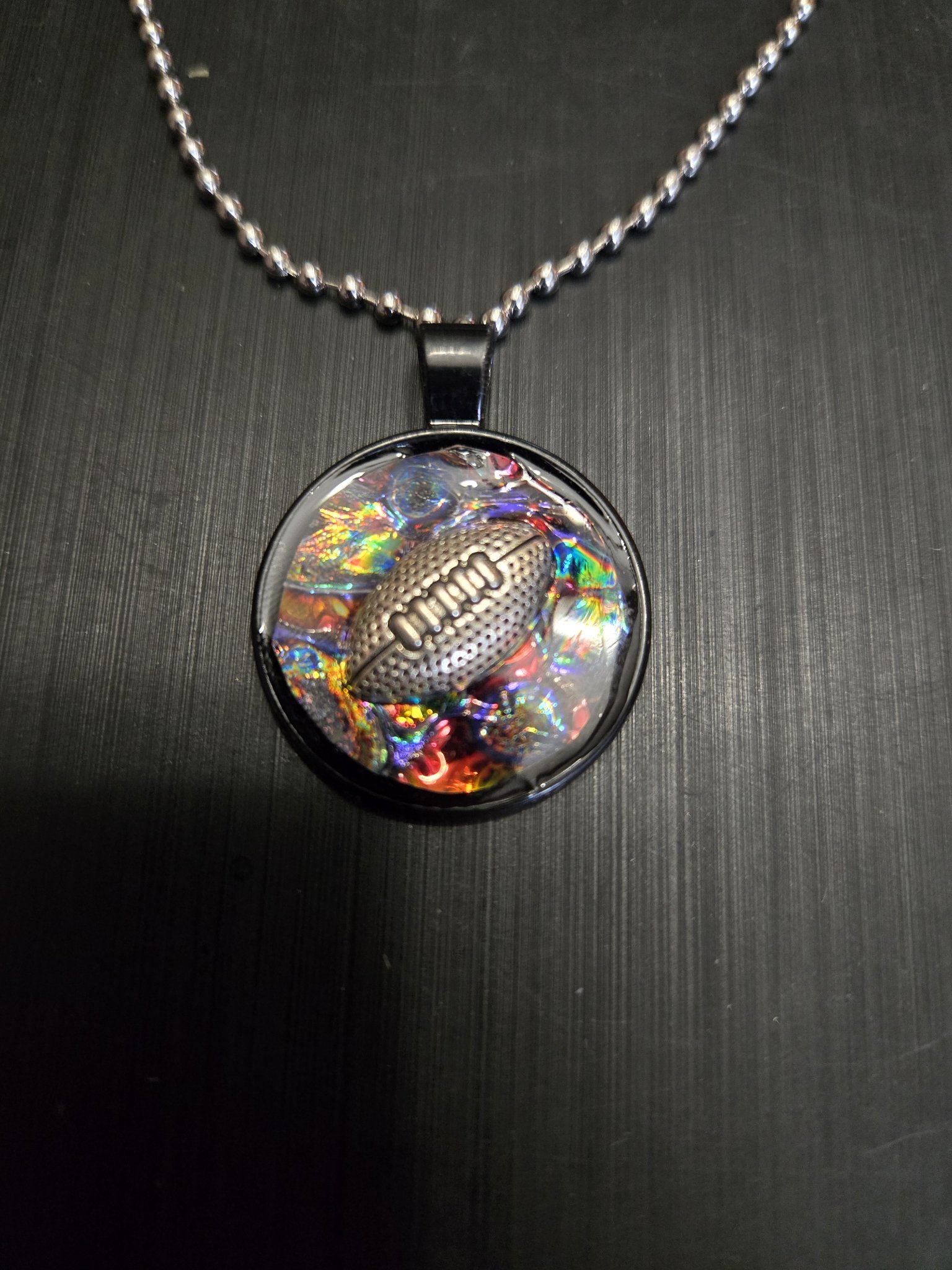 Front view of the round rainbow holographic football pendant with a silver-tone football charm in a black metal frame on its 22-inch ball chain