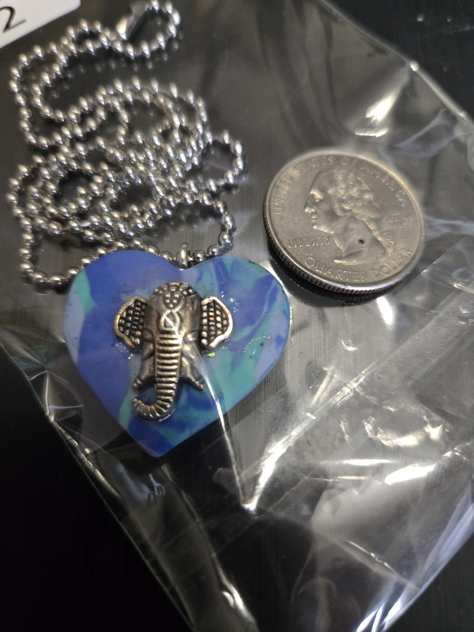 Front of the blue and teal heart elephant pendant beside a quarter showing it is approximately two inches across