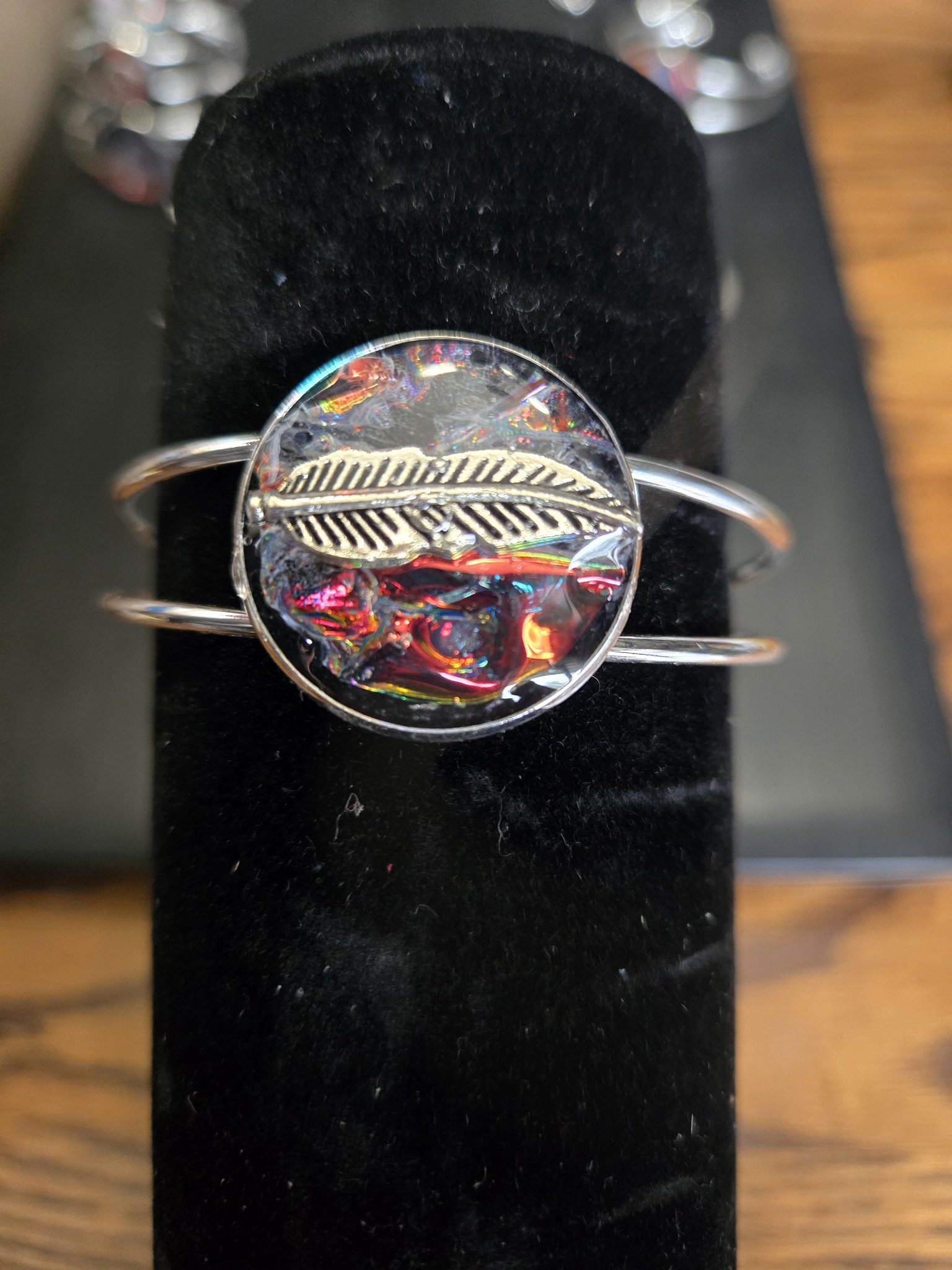 Silver-tone adjustable cuff bracelet on a black display stand with a feather over a colorful holographic resin medallion