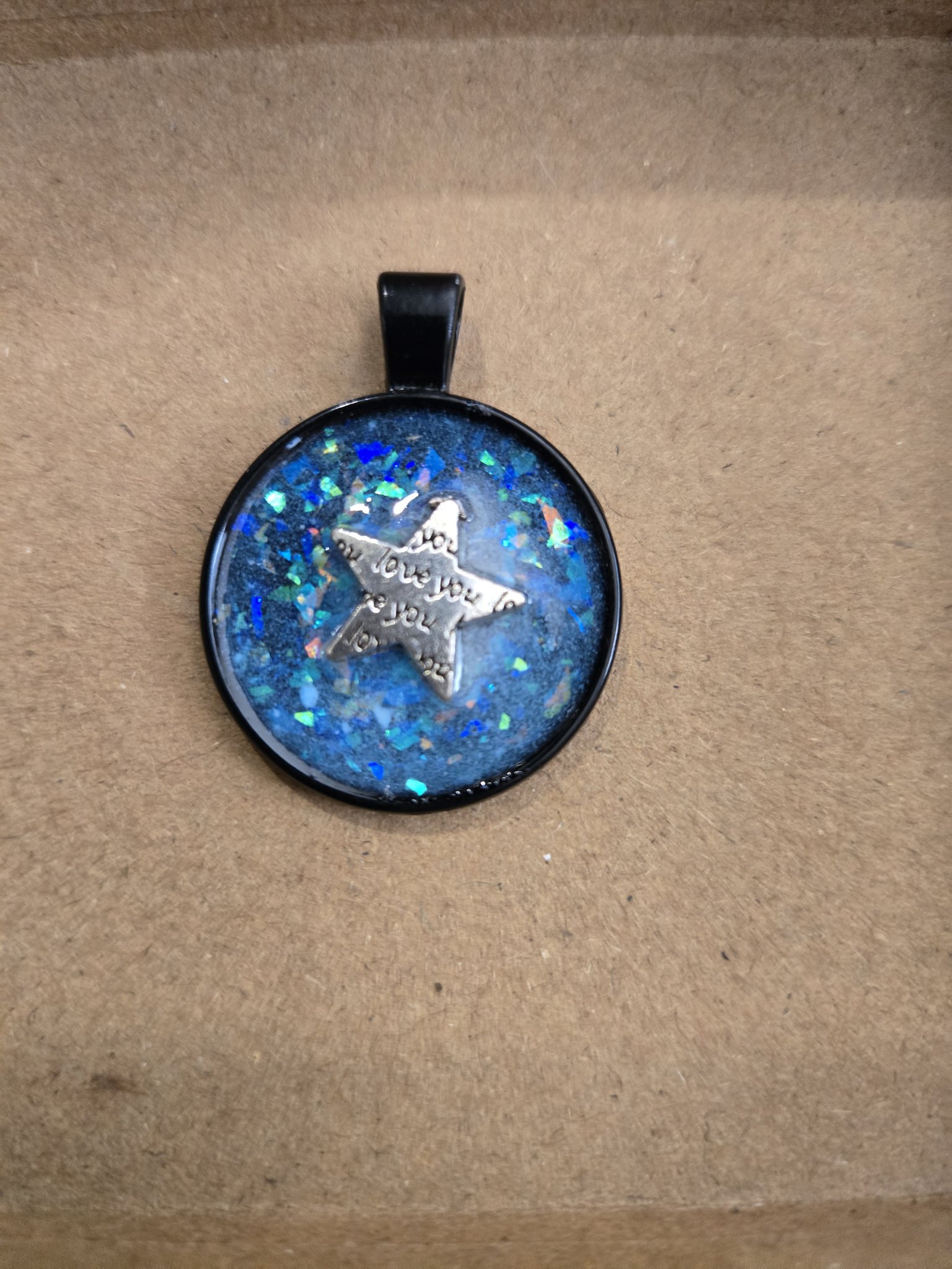 Stock 169 round blue holographic glitter resin pendant in a black metal frame with a silver-tone love you star charm in daylight