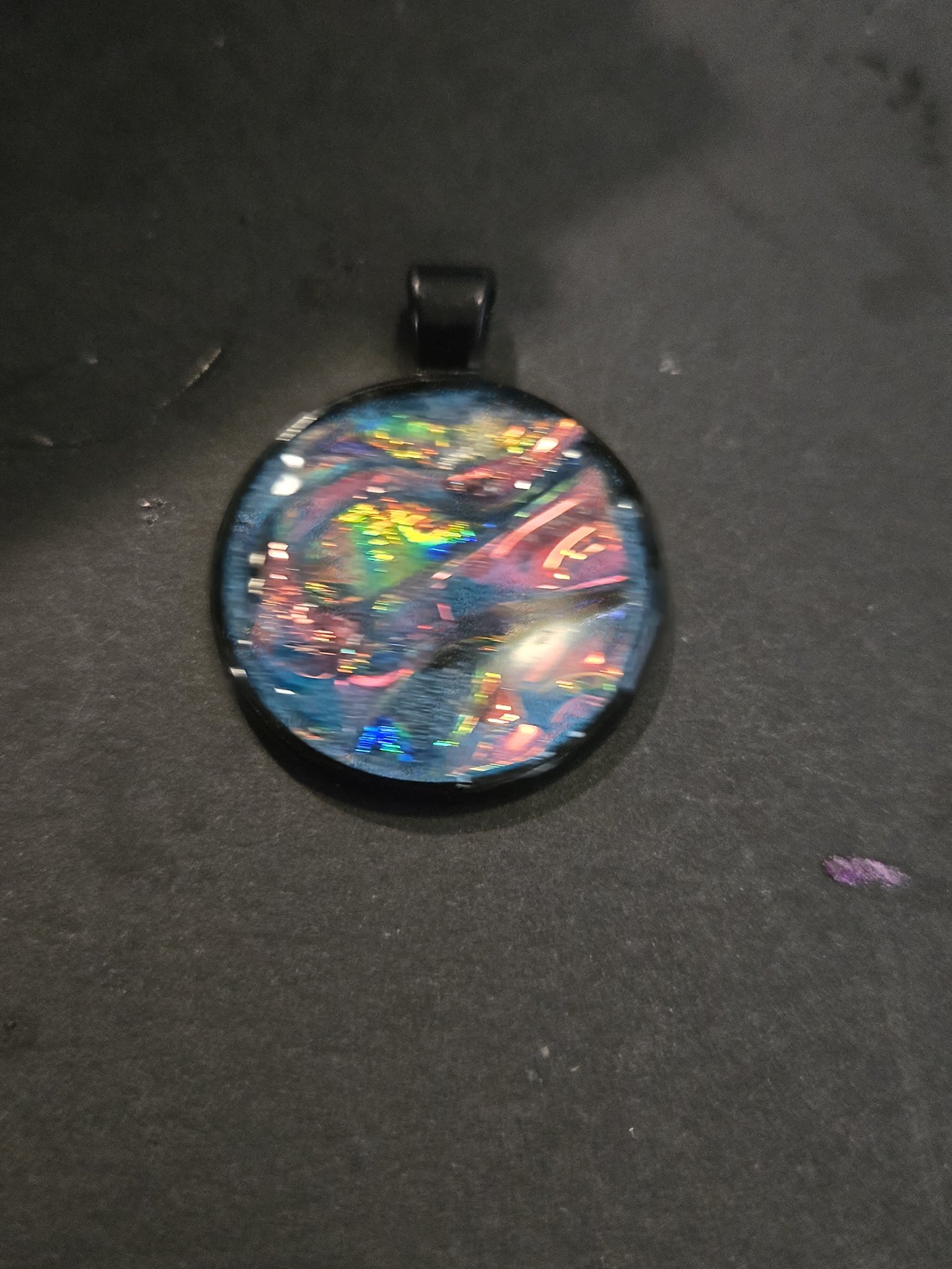 Stock 204 round blue glow-in-the-dark holographic resin pendant with rainbow swirls in a black metal frame, shown from the front