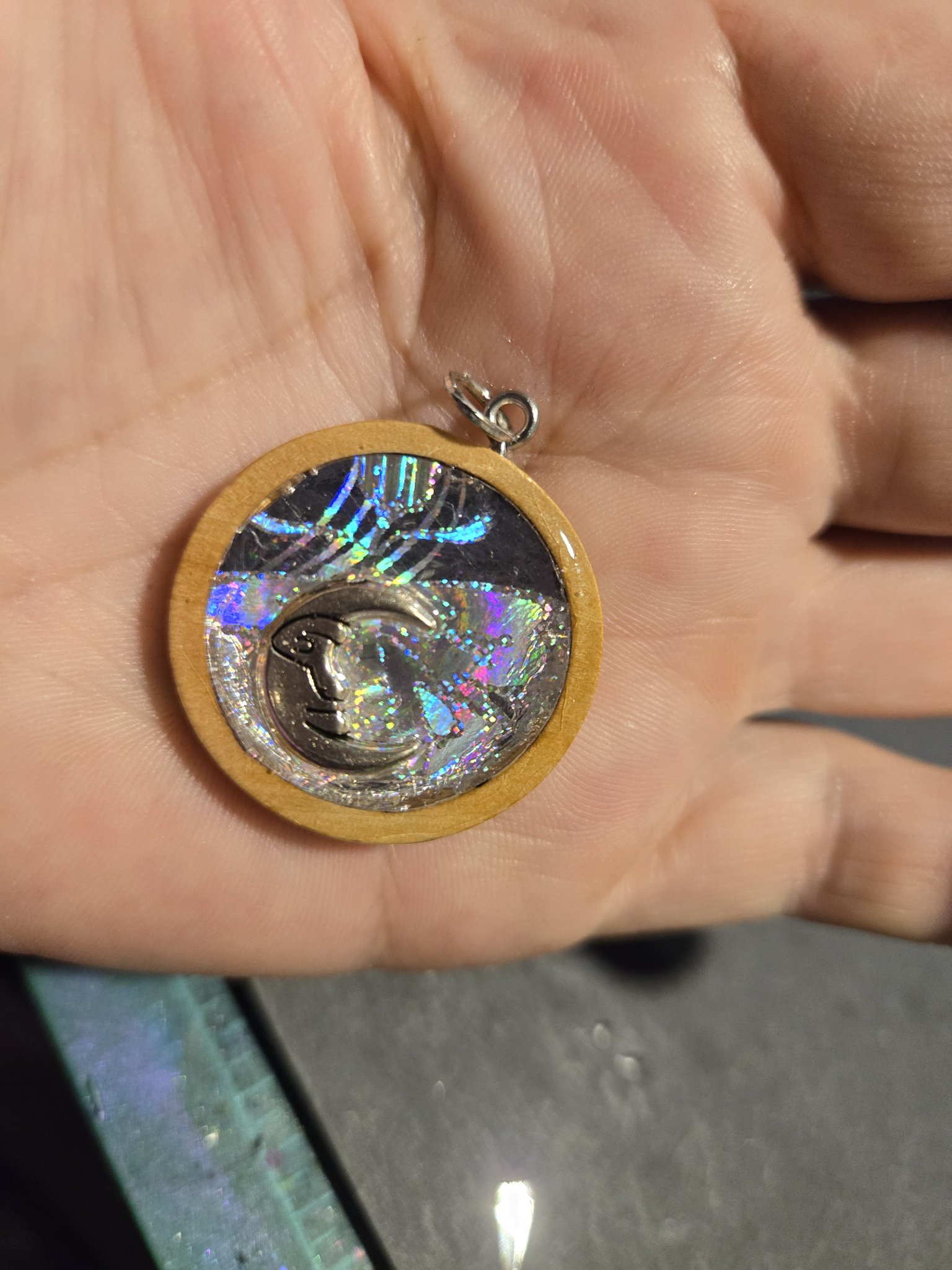 Stock 209 round holographic resin pendant on a wood backing with a silver-tone crescent moon charm, shown from the front in hand