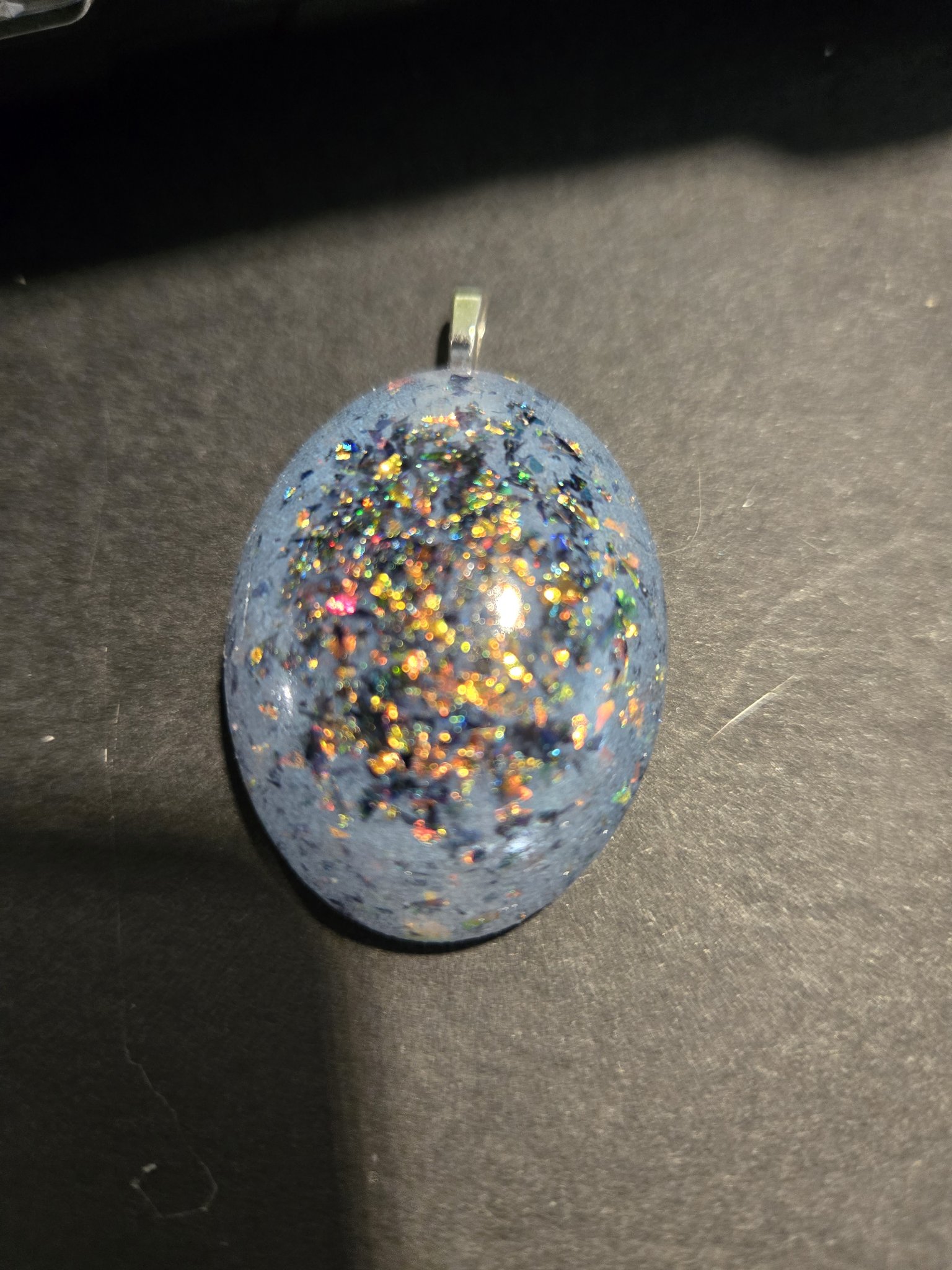 Stock 251 holographic glow oval pendant on a dark background, showing the soft blue resin scattered with rainbow holographic flecks