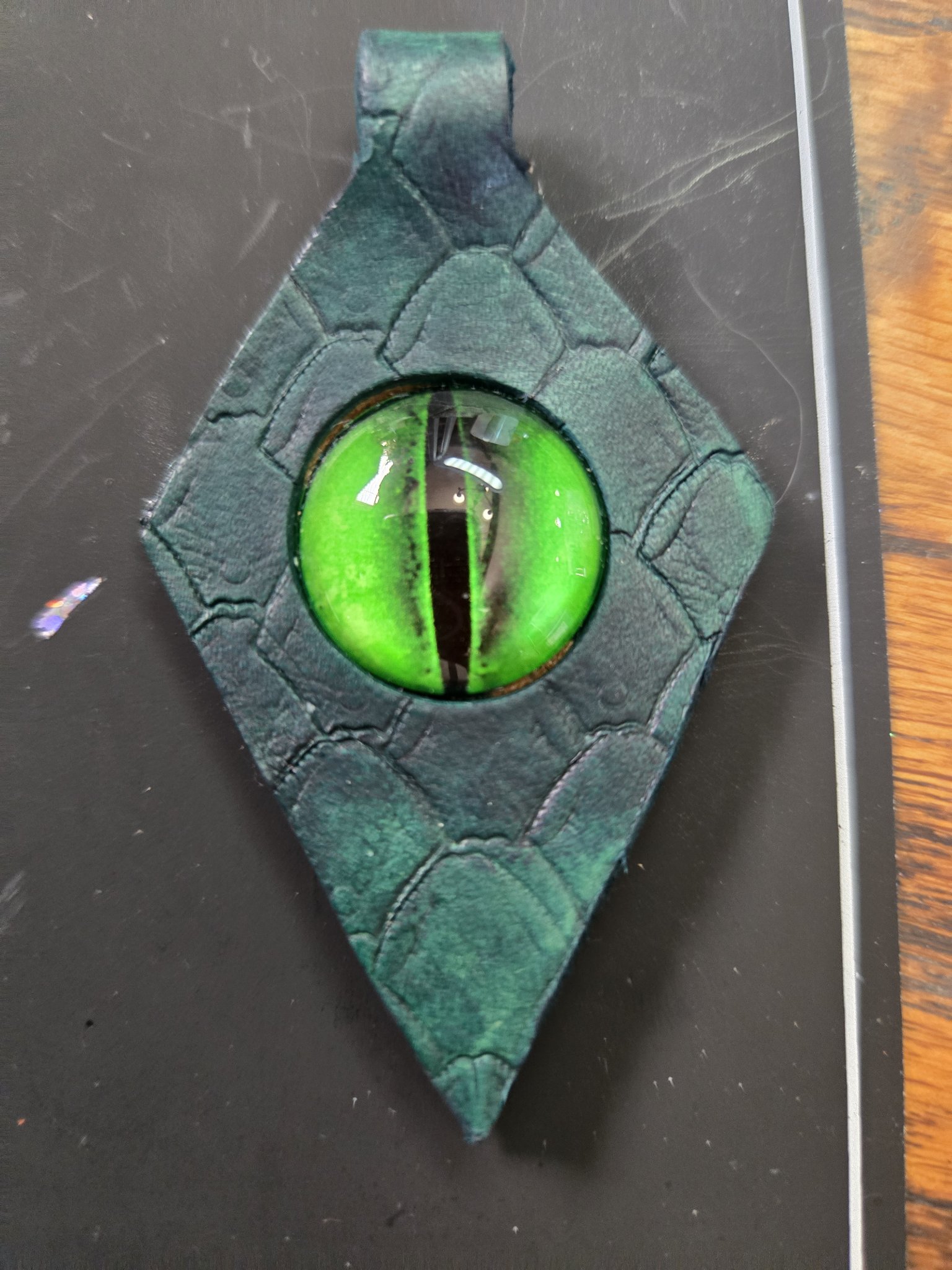 Front view of a black leather pendant with stamped dragon-scale texture and a bright green dragon eye glass cabochon