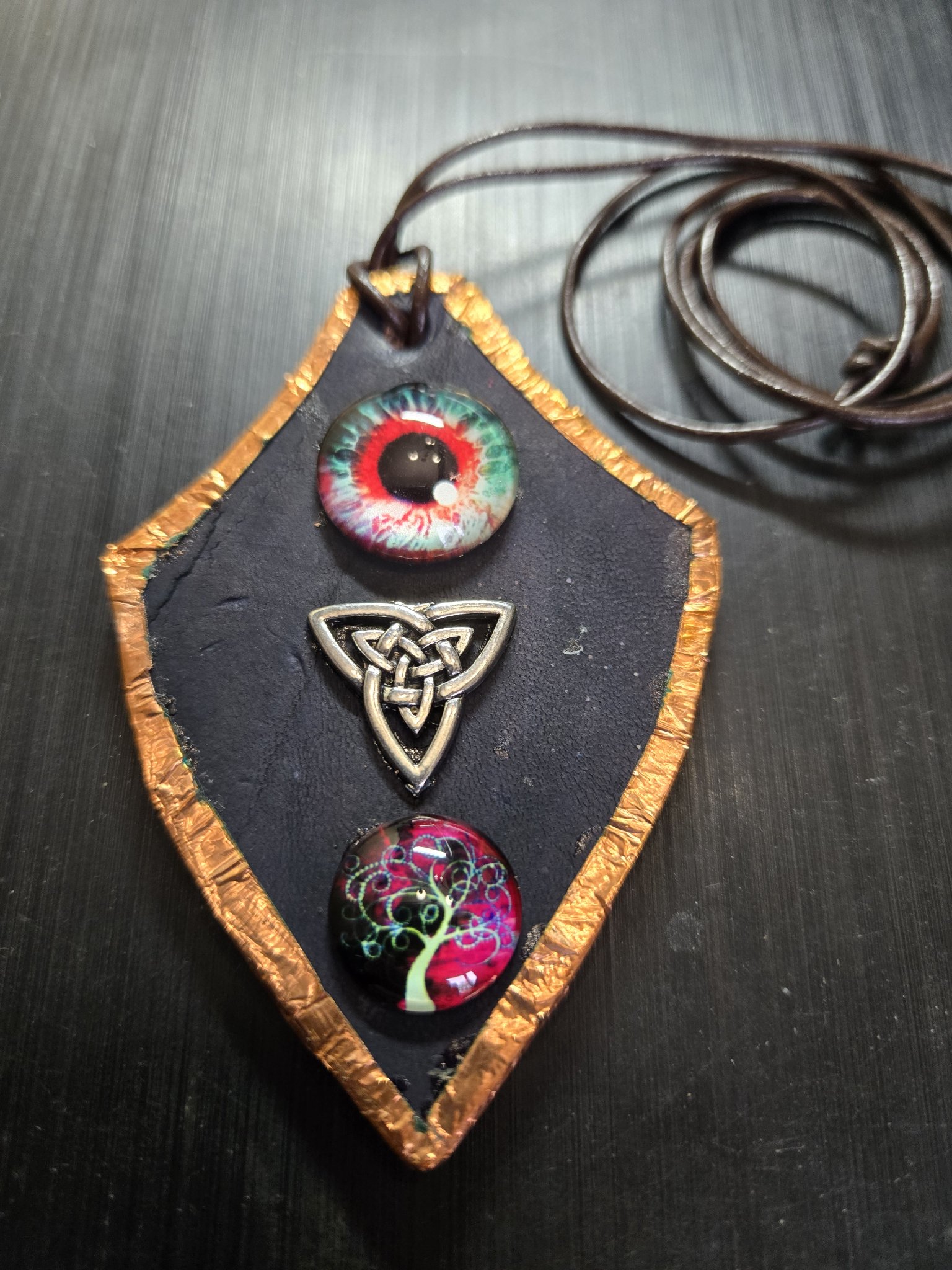 Front close-up of stock 161, a shield-shaped black leather pendant framed in copper with a Celtic triquetra knot, glass eyeball cabochon, and glass Tree of Life cabochon