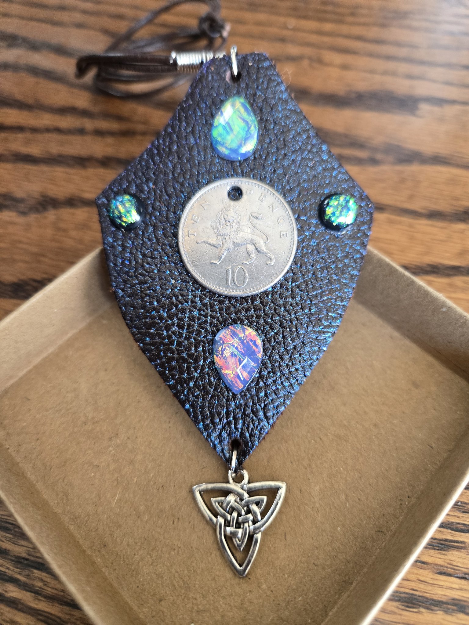 Stock 175 buffalo leather shield pendant with blue mica sheen, a British 10 pence lion coin, holographic cabochons, and a silver-tone Celtic triquetra charm, shown in a front close-up