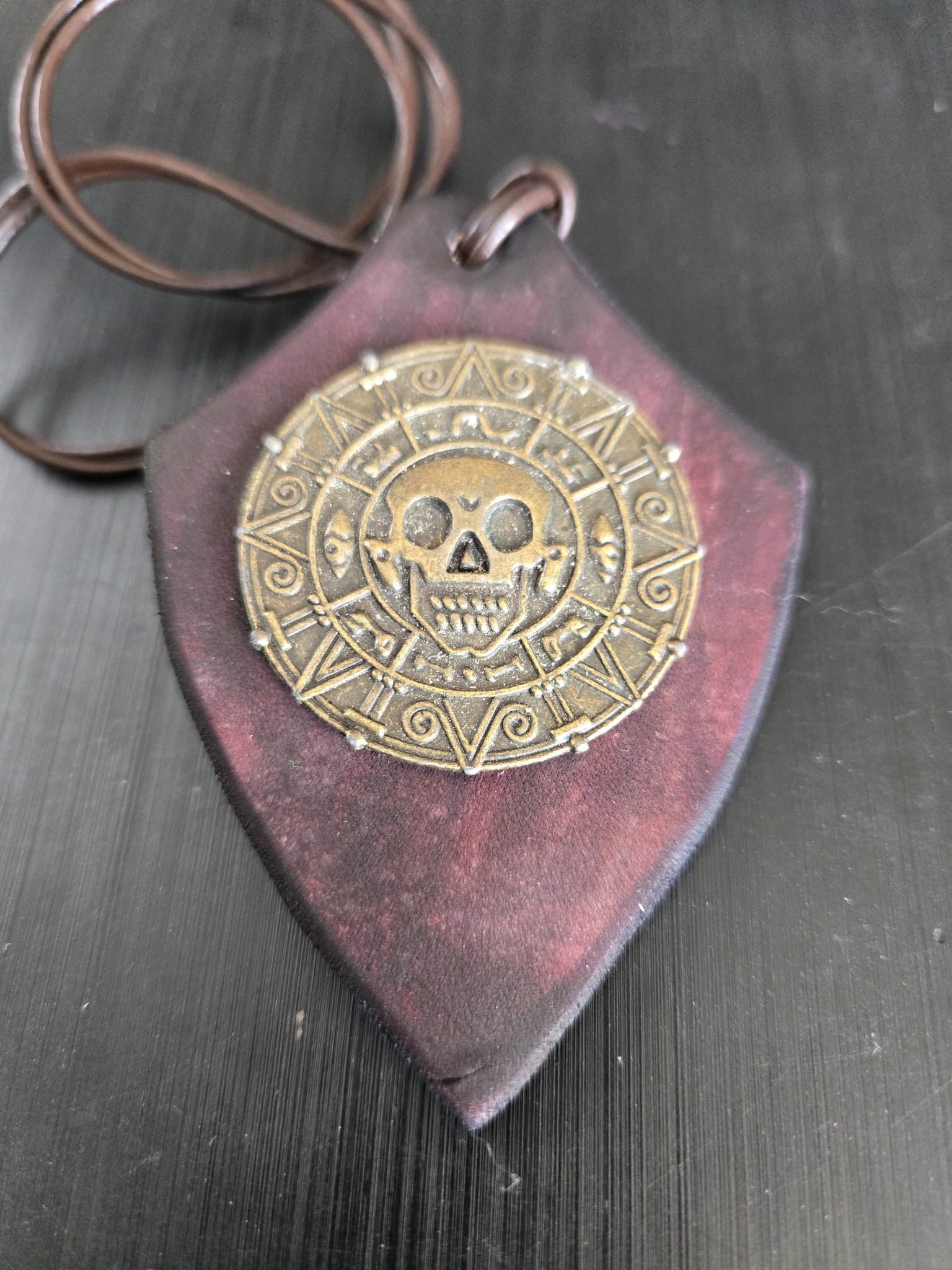 Stock 179 shield-shaped dark maroon leather pendant with a brass-tone Aztec-style skull coin medallion, shown from the front with its brown leather cord