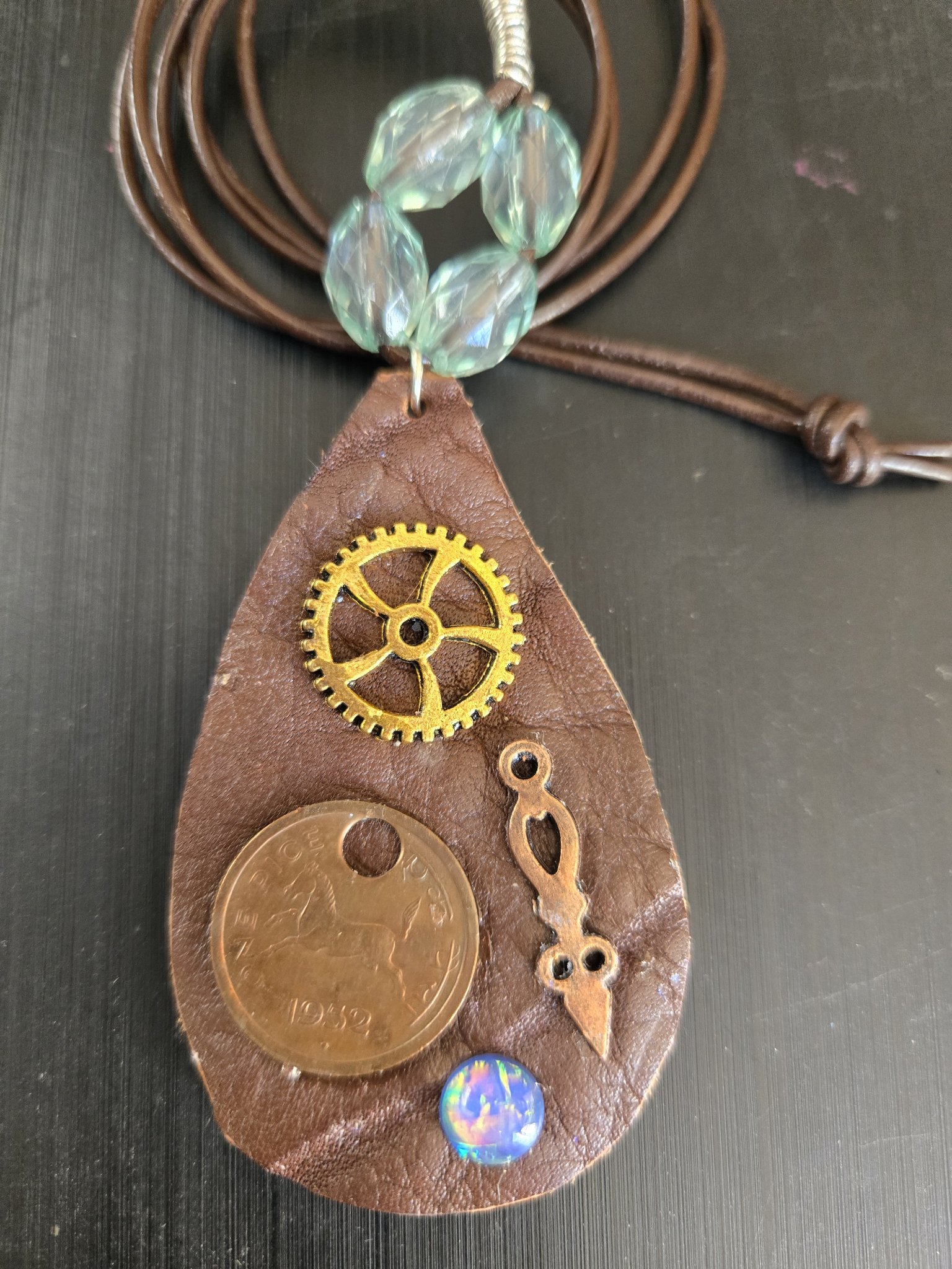 Stock 180 teardrop brown buffalo leather pendant with a gold-tone gear, 1952 one pice coin, copper clock hand, blue holographic cabochon, and faceted pale blue beads at the bail, shown in a front close-up