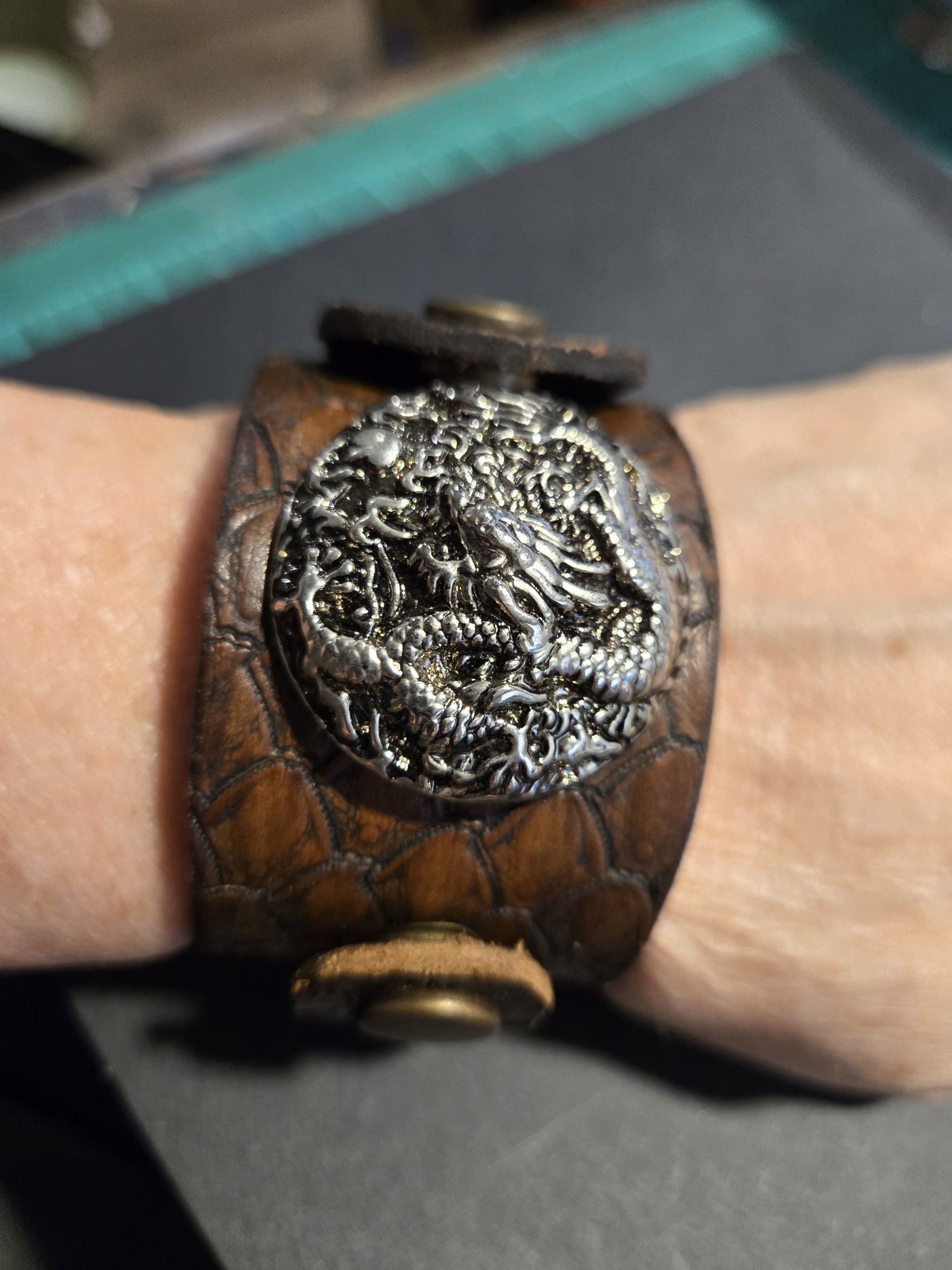 Stock 214 brown leather cuff with hand-stamped dragon scales, a silver-tone dragon emblem, and bronze snap closure, shown on a wrist